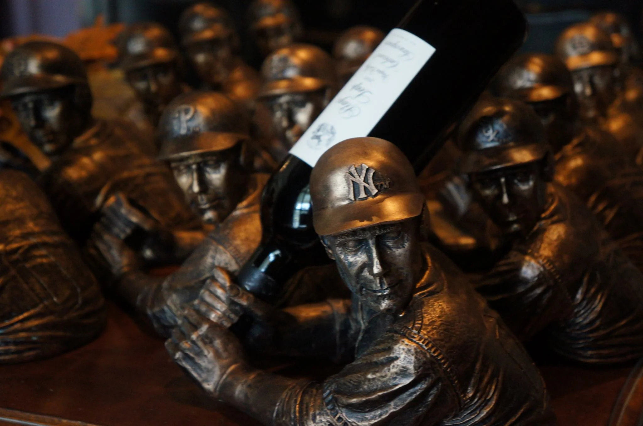 A row of original BamVino bottle holders in bronze, wearing MLB team helmets — Yankees, Phillies and others